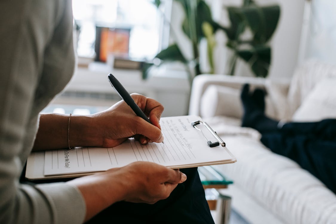 Free Unrecognizable ethnic female therapist taking notes on clipboard while filling out form during psychological appointment with anonymous client lying on blurred background Stock Photo